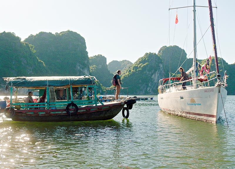 Checker Tobi steht auf dem Deck eines vietnamesischen Bootes und begrüßt Marina auf dem Deck eines kleinen weißen Segelschiffs
