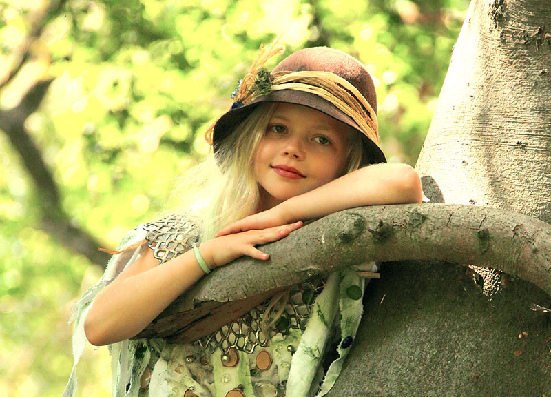 Lene trägt einen Hut und lehnt sich verträumt an einen Baum