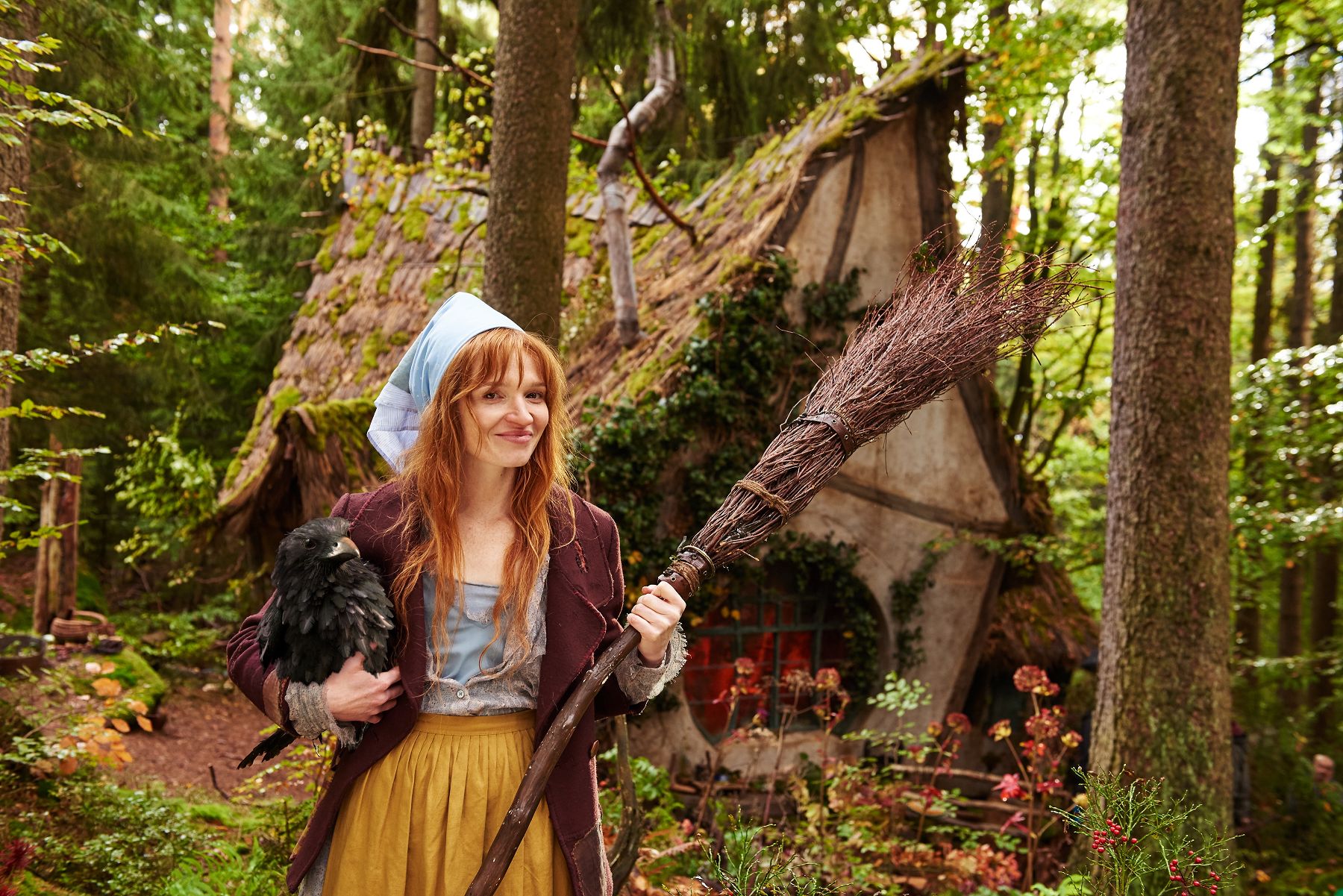 Die kleine Hexe mit rotem Haar und Kopftuch steht lächelnd mit ihrem Besen und einem schwarzen Raben im Wald vor ihrem windschiefen, moosbedeckten Hexenhaus.
