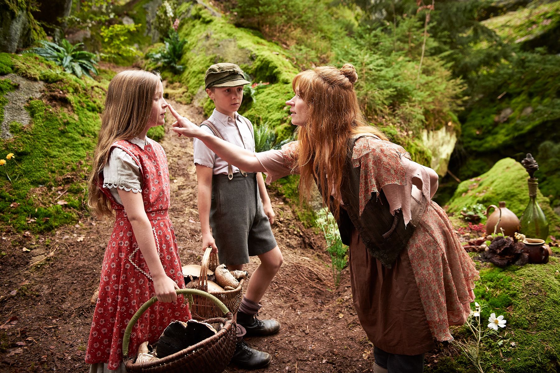 Die kleine Hexe beugt sich im Wald zu zwei Kindern hinab und tippt dem Mädchen spielerisch auf die Nase. Der Junge daneben trägt einen geflochtenen Korb mit Pilzen
