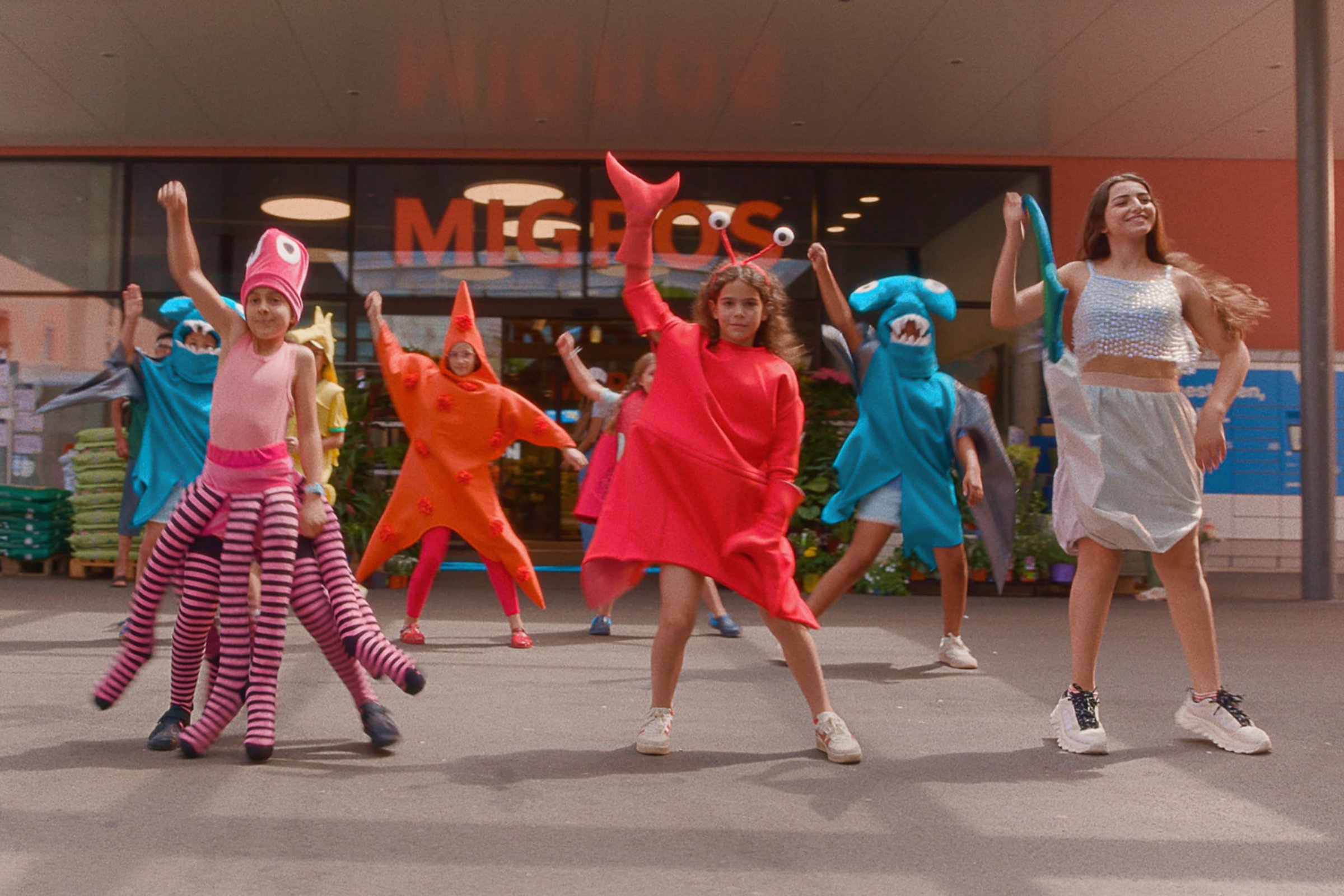 Eine Gruppe von Kindern tanzt in selbstgebastelten Meereskostümen, darunter ein roter Krebs, eine Krake, ein Seestern und ein Hai, auf dem Vorplatz eines Supermarkts.