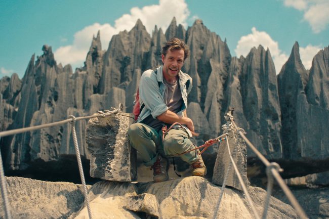 Tobi hockt an einer Seilbrücke vor spitzen Bergen