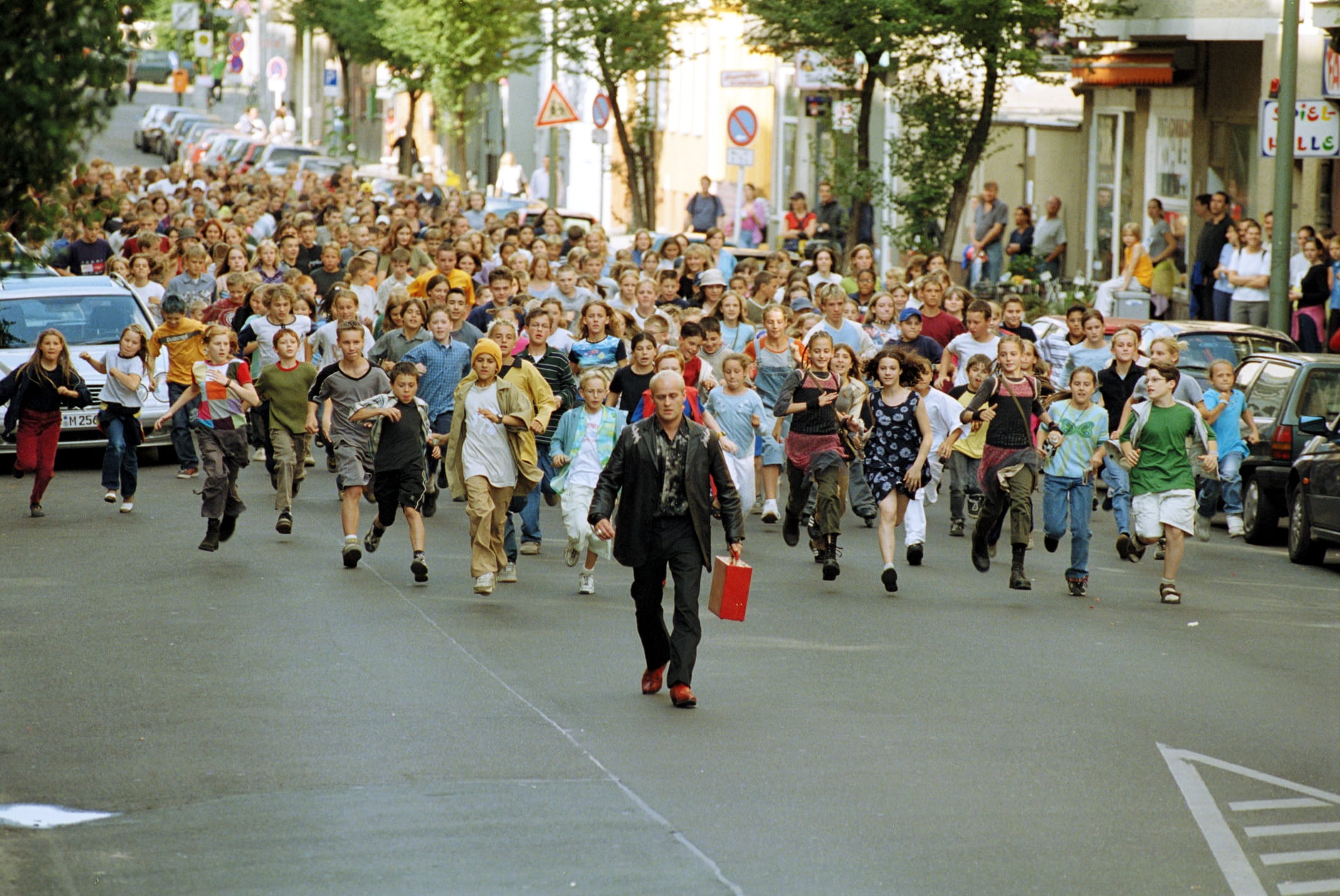 Emil und eine riesige Menge rennender Kinder verfolgen Max Grundeis, der mit seinem roten Koffer über eine Berliner Straße flüchtet.