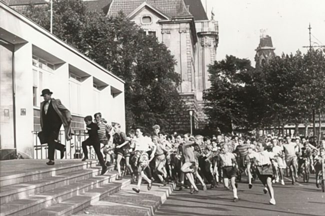 Eine große Gruppe von Kindern jagt im Laufschritt einen flüchtenden Mann mit Hut und hellem Mantel eine Treppe in der Stadt hinunter.