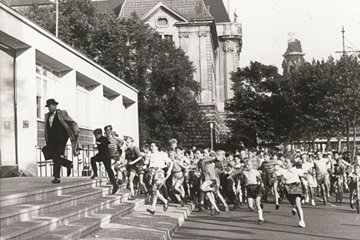 Eine große Gruppe von Kindern jagt im Laufschritt einen flüchtenden Mann mit Hut und hellem Mantel eine Treppe in der Stadt hinunter.