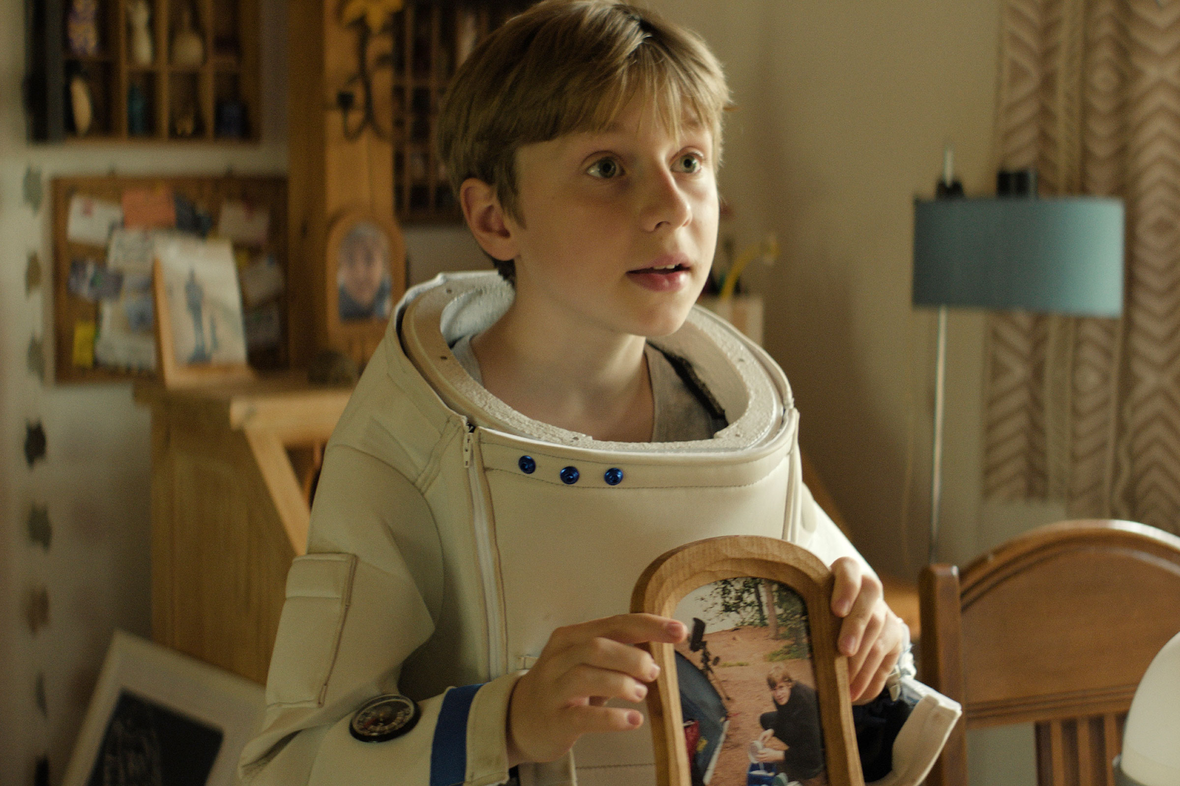 Tom steht in seinem Astronautenanzug, aber ohne Helm, im Zimmer und hält fragend ein Bild