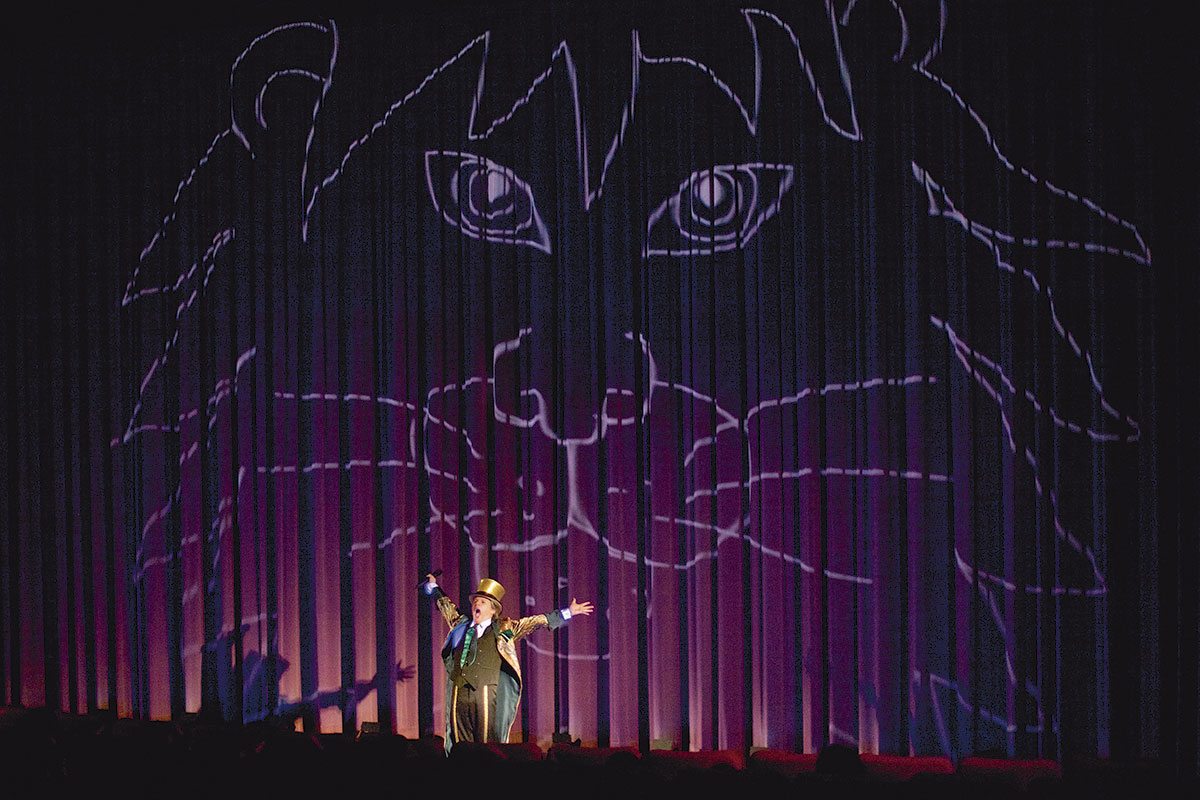 Marquinhos im Kino auf der Bühne breitet die Arme aus, steht vor einem Vorhang auf dem der Theo-Kopf als Projektion zu sehen ist