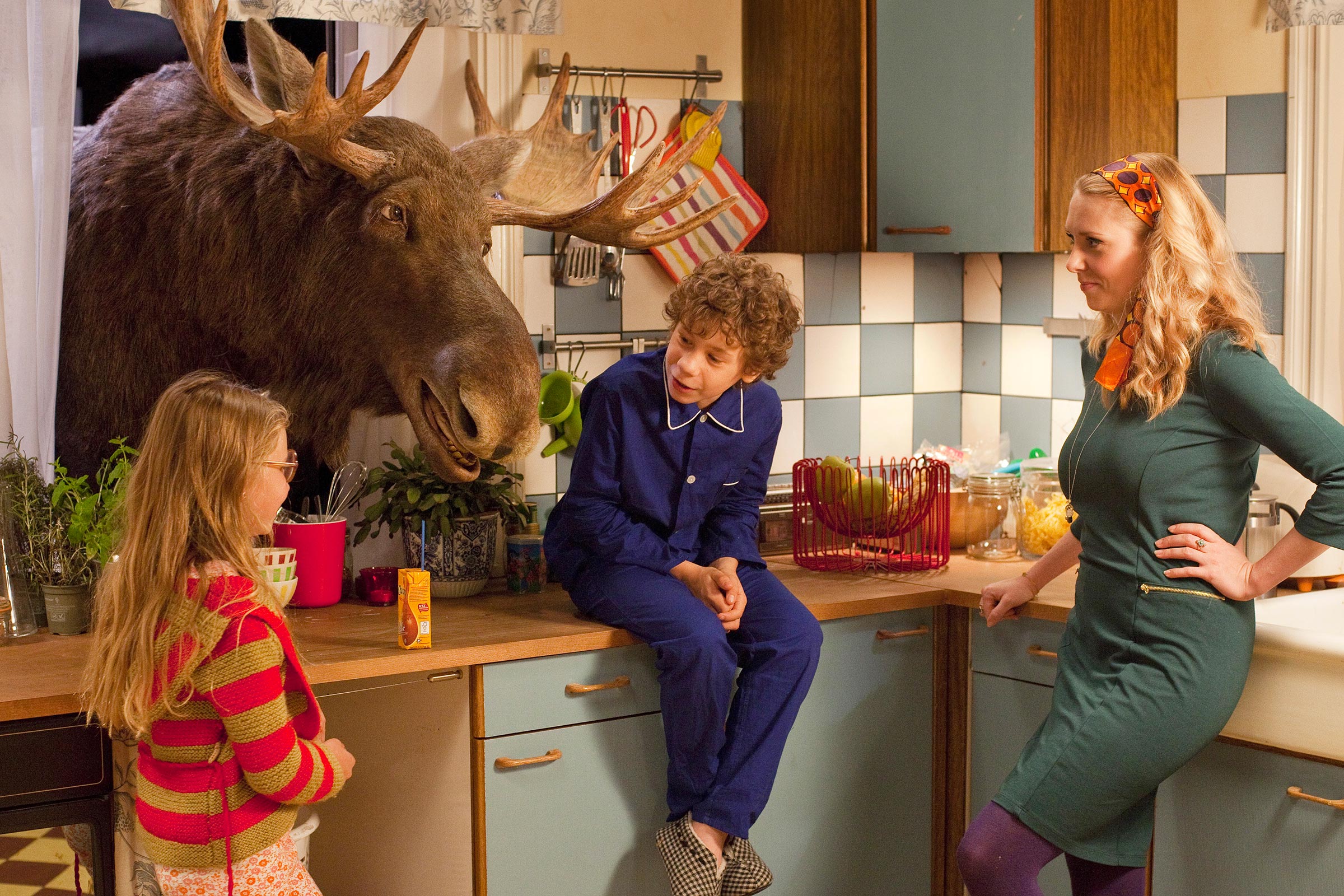 Max mit seiner Schwester und seiner Mutter in der Küche, der Elch streckt seinen Kopf durch das geöffnete Fenster und redet mit