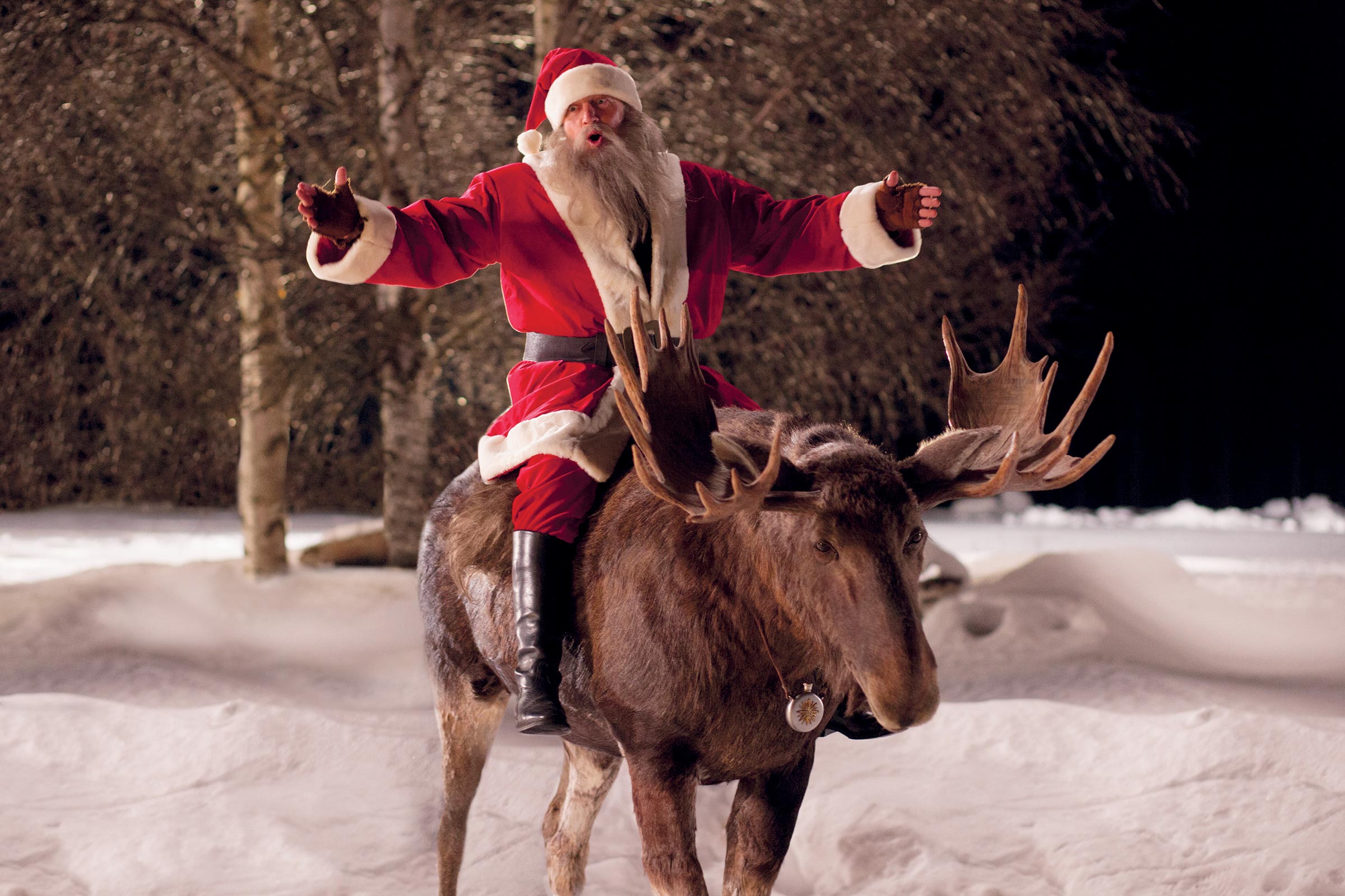Der Weihnachtsmann reitet mit weit ausgebreiteten Armen auf einem Elch durch die Schneelandschaft