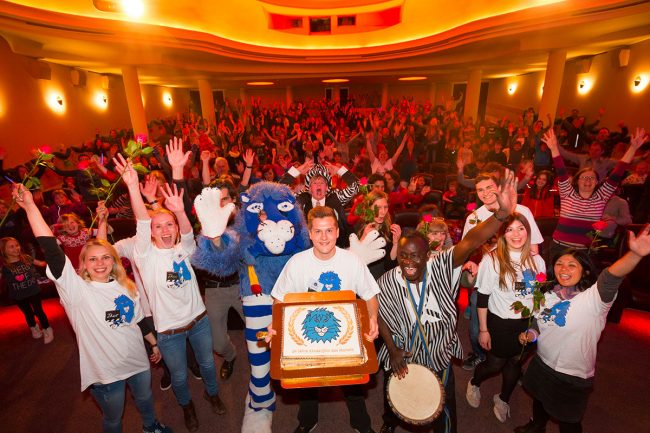 Eine Gruppe aus Helferinnen und Helfern steht mit Theo und einem Musiker mit einer Geburtstagstorte vor dem Publikum und alle jubeln und recken die Hände hoch