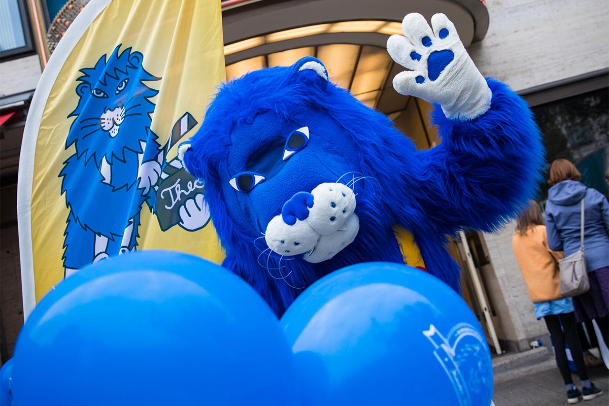 Theo steht vor dem Kino und hebt die Hand zum Gruß, im Vordergrund zwei blaue Luftballons