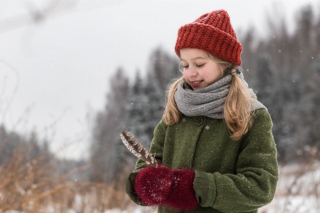 Eia steht in der verschneiten Landschaft, hält eine Feder in der Hand und betrachtet sie lächelnd